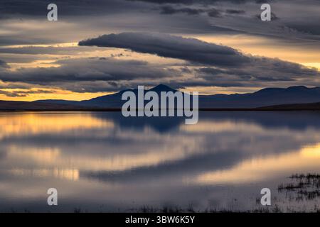 Blick auf das stille, reflektierende Wasser, das den dramatischen Himmel und die fernen Berge unter einem sanften, goldenen Licht spiegelt, Island. Stockfoto