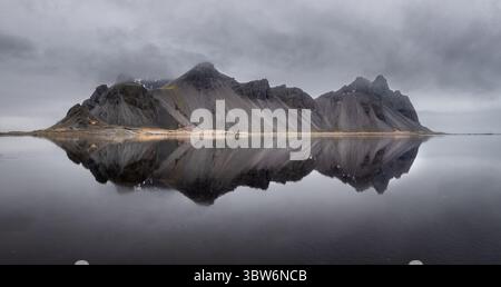 Blick auf schroffe, zerklüftete Berge, die sich im stillen, dunklen Wasser unter einem brodelnden Himmel spiegeln, eine ätherische Landschaft, Hofn, Island. Stockfoto
