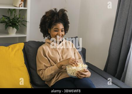 Afroamerikanische Frau sitzt auf einem grauen Sofa und geniesst eine Schuessel Popcorn, waehrend sie einen Film oder einen Online-Streaming-Film Ansie Stockfoto