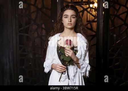 Eine junge schöne Brünette mit langen losen Haaren in einem weißen Peignoir in der Mode des 19. Jahrhunderts hält eine scharlachrote Rose in den Händen - ein Cosplay f Stockfoto