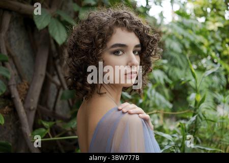 Frau mit braunen Haaren und blauem Kleid. Sie steht in einem grünen Wald. Sie trägt eine Halskette Stockfoto