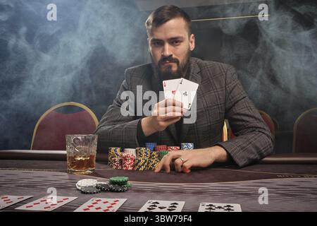 Ein bärtiger Mann in einem grauen Anzug spielt Poker und sitzt im Rauch am Tisch im Casino. Er zeigt die Siegerkombination aus zwei Assen in seiner Hand A Stockfoto