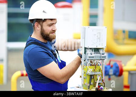 Ein Ingenieur in einem Helm und Arbeitskleidung passt die Ausrüstung in der Produktionsstätte an und inspiziert sie. Ein authentisches Porträt eines kaukasischen Mannes mit Stockfoto