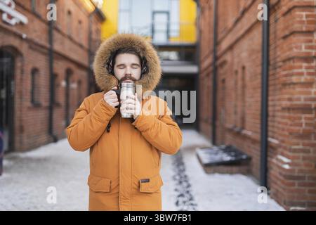 Nahaufnahme Porträt hübscher bärtiger tausendjähriger Mann in Winterkleidung und mit Thermosschnee draußen. Kalte Jahreszeit und heißes Getränk im Winter Stockfoto