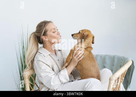 Eine junge, schöne Frau in lässiger Kleidung umarmt und streichelt ihren geliebten Hund, der im Schlafzimmer ihres gemütlichen Landhauses sitzt. Tierisches Kommunikationskonzept Stockfoto