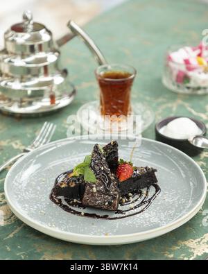 Schokoladen-Brownie-Scheiben werden auf einem Teller mit Schokoladensoße, Erdbeere, Nüssen, Puderzucker und Minze mit einem Glas Tee und türke serviert Stockfoto