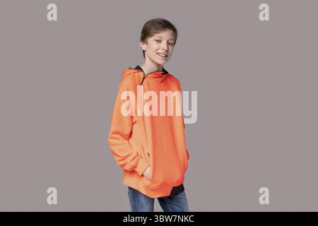 Porträt glücklicher kaukasischer Teenager-Mann in orangefarbenem Sweatshirt auf grauem Hintergrund. Fröhlich lächelndes kaukasisches männliches Model mit einem weißen Lächeln in einer Stange Stockfoto