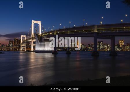 Beleuchtete Regenbogenbrücke bei Nacht in Tokio, Japan. Skyline der Stadt und Tokio Tower im Hintergrund sichtbar. Atemberaubender Blick auf die Stadt in der Dämmerung. Stockfoto