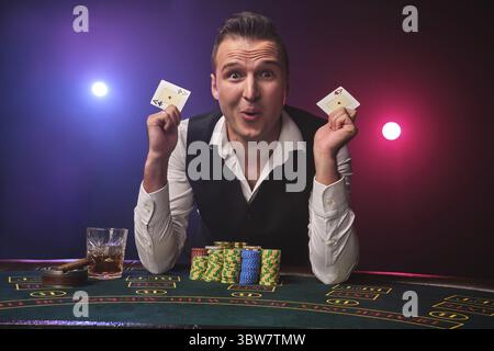 Ein gutaussehender reifer Mann in weißem Hemd und schwarzer Weste hält zwei Asse in der Hand, um seinen Sieg zu feiern. Er spielt Poker mit einer Aufregung Stockfoto