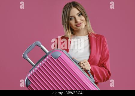 Charmante blonde Frau in rotem Strumpfanzug und weißer Bluse. Sie lächelt und hält ihren Koffer, während sie vor rosafarbenem Hintergrund posiert. Mode, Reisen Stockfoto