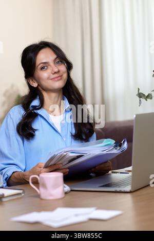 Porträt der Frau in blauem Hemd hält Ordner verwendet Laptop im Heimbüro Stockfoto