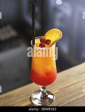 Farbenfroher Cocktail steht auf einer hölzernen Bartheke mit schwarzem Stroh, Orangenscheibe und Himbeeren Stockfoto