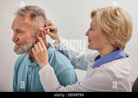 Erfahrene Ärztin, die hinter-dem-Ohr-Hörgerät an das Ohr des älteren Patienten anpasst, während der Sprechstunde in der audiologischen Klinik Stockfoto