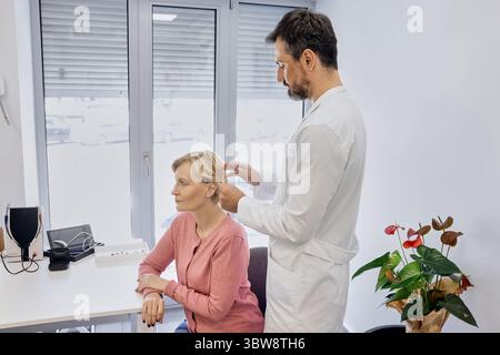 Erfahrener männlicher Arzt, der hinter-dem-Ohr-Hörgerät an das Ohr des älteren Patienten anpasst, während der Sprechstunde in der audiologischen Klinik Stockfoto