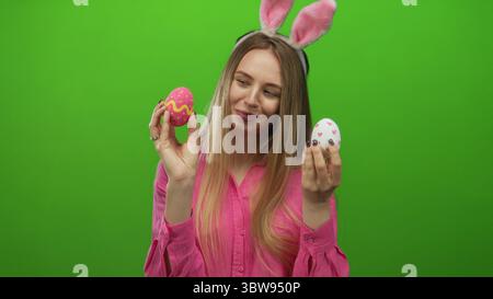 Junge Frau mit Hasenohren hält ostereier vor einem hellgrünen Hintergrund und lächelt fröhlich über farbenfrohe Dekorationen in festlicher Kleidung. Stockfoto