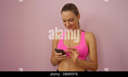 Frau lächelt beim Verwenden des Smartphones in rosa Sportoberteil vor isoliertem rosa Hintergrund und vermittelt Fitness und Konnektivität. Stockfoto
