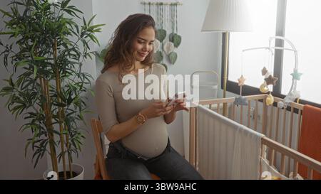 Schwangere Frau lächelt und macht ein Selfie in einem gemütlichen Schlafzimmer, das mit einer Holzkrippe und grünen Pflanzen geschmückt ist, die Glück und Vorfreude in einer se verkörpern Stockfoto