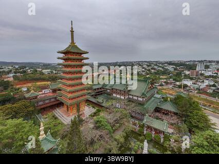 Aus der Vogelperspektive auf den majestätischen Turm der Minh Thanh Pagode, der die Skyline inmitten der Umarmung der Stadt durchdringt, eine Symphonie aus Grün und Gold unter einem ruhigen Himmel, Pleiku, Gia Lai, Vietnam. Stockfoto