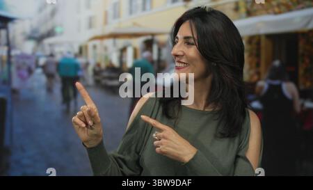 Die hispanische Frau lächelt und zeigt mit dem Finger auf das Objekt auf der Außenterrasse des Restaurants; freundliche Einladung. Stockfoto
