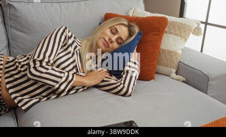Frau mit gestreiftem Pyjamaoberteil, die im Studio auf einem grauen Sofa liegt, mit geschlossenen Augen und entspannter Haltung; Ruhe. Stockfoto
