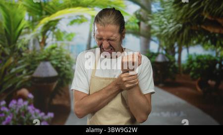 Mann trägt beige Schürzengrimaces, während er das Handgelenk auf einem von tropischem Grün gesäumten Parkweg hält; Unwohlsein. Stockfoto