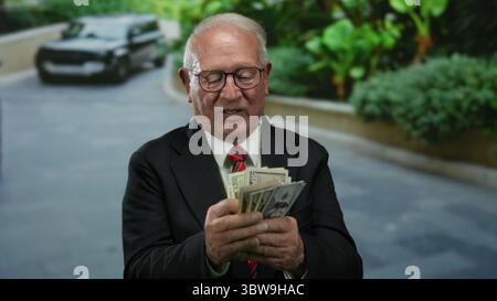 Ein Senior-Geschäftsmann zählt uns im Freien Dollar mit einem Luxusauto im Hintergrund, der Reichtum und Erfolg in einer modernen urbanen Umgebung demonstriert. Stockfoto