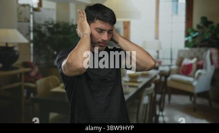 Der Mann bedeckt die Ohren mit beiden Händen zwischen gemütlichen Wohnzimmerstühlen und Lampe in einem Gebäudeinneren; Distress. Stockfoto
