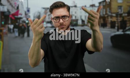 Junger bärtiger Mann auf einer Stadtstraße, der eine unhöfliche Handbewegung mit beiden Mittelfingern macht, während er Brille und ein schwarzes Hemd in einem urbanen Outd trägt Stockfoto