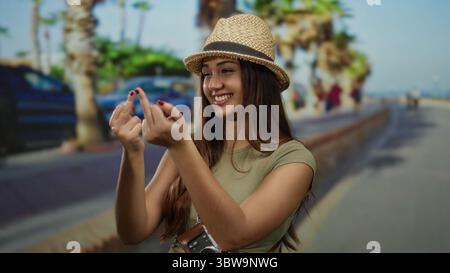 Frau lächelt draußen und macht Gesten in der sonnigen Straße mit Palmen, die Hut und lässige Kleidung tragen und während des Urlaubs verspielte Touristenmanieren zeigen. Stockfoto