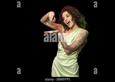 Glücksspiel begeisterte junge Brünette mit Tattoos auf den Armen, die vor schwarzem Hintergrund stehen, lachen, während sie einen Stapel von Wettchips in den Händen hält, Croppe Stockfoto