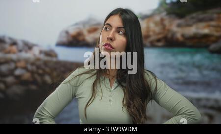 Junge hispanische Frau, die am Strand steht und nachdenklich in Richtung des ruhigen Ozeans unter klarem Himmel blickt und eine natürliche kontemplative Stimmung zeigt Stockfoto