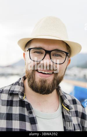 Nahaufnahme schöner Millennials Mann mit Hut und Brille in der Nähe des Yachthafens mit Yachten. Porträt eines lachenden Mannes mit Seehafenhintergrund Stockfoto