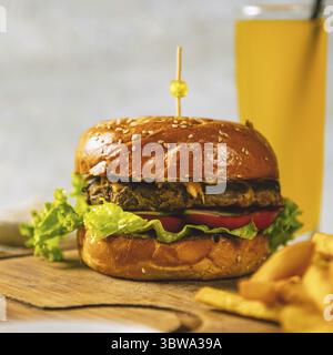 Saftiger Burger steht auf einem Holzbrett mit Pommes Frites und einem Glas Orangensaft im Hintergrund Stockfoto