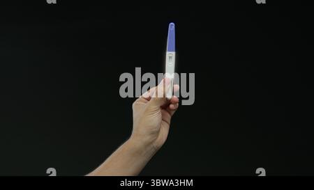 Mann hält Schwangerschaftstest über schwarzem Hintergrund, Hand isoliert, zeigt Ergebnis in hellem Raum, erfasst einen intimen und signifikanten Moment in Heilung Stockfoto