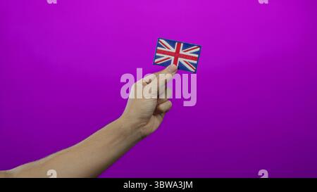 Die männliche Hand hält einen gestickten unions-Jack vor rosa Hintergrund, der die Identität großbritanniens und den kulturellen Stolz symbolisiert, isoliert mit lebhafter Betonung. Stockfoto