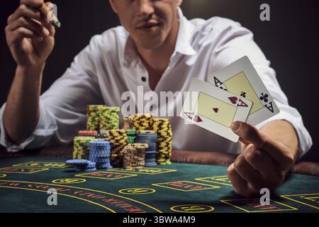 Ein sportlicher Mann in einem weißen Hemd spielt Poker und sitzt im Rauch am Tisch im Casino. Er freute sich über seinen Sieg und zeigte zwei Asse in der Hand. Gambli Stockfoto