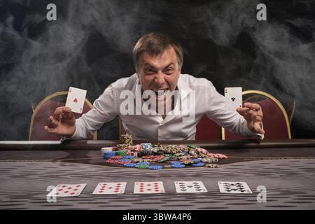 Ein ausdrucksstarker Kerl in einem weißen Hemd spielt Poker und sitzt im Rauch am Tisch im Casino. Er freut sich, zwei Asse in der Hand zu zeigen Stockfoto