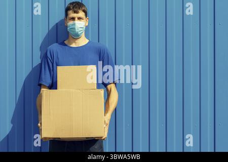 Ein junger kaukasischer Kurier in einer medizinischen Maske hält Pappschachteln in der Hand. Das Konzept der Paketzustellung oder der humanitären Hilfe Stockfoto