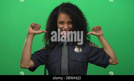 Hispanische Offizierin in Uniform lächelt selbstbewusst mit Daumen, die vor einer grünen Hintergrundwand auf sich gerichtet sind. Stockfoto