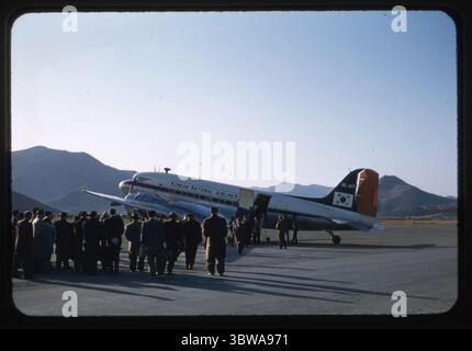 Ein seltenes Farbfoto einer Korean National Airlines Douglas DC-3 (Kennzeichen HL-007) als Passagiere am K-9 Airfield in Pusan (Busan), Südkorea, im Dezember 1956. Dieses Bild fängt die frühe Ära der südkoreanischen Zivilluftfahrt ein, wobei die Lackierung der Fluggesellschaft und das Taegukgi-Logo deutlich sichtbar sind. Die Korean National Airlines (KNA) wurde 1946 gegründet und war die erste kommerzielle Fluggesellschaft Südkoreas, die in der Nachkriegszeit DC-3-Flugzeuge Betrieb. Die Kleidung der Menschenmenge und die minimalistische Infrastruktur des Flugplatzes spiegeln die Übergangsphase Koreas zwischen Konflikt und Modernisierung wider. Stockfoto