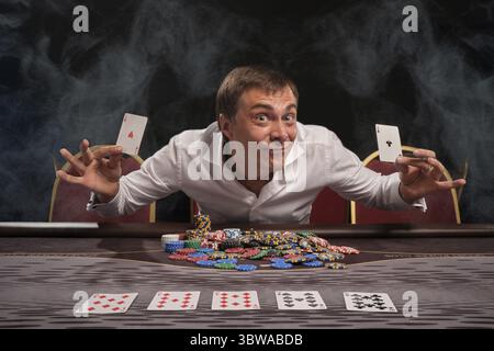 Ein ausdrucksstarker Mann in einem weißen Hemd spielt Poker und sitzt im Rauch am Tisch im Casino. Er freut sich, zwei Asse in der Hand zu zeigen. Gambli Stockfoto