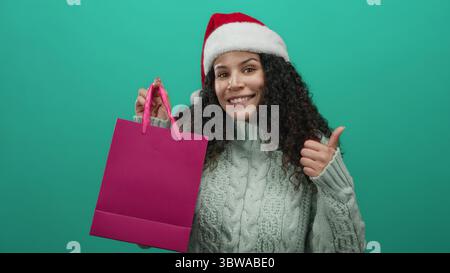 Glückliche junge hispanische Frau mit weihnachtsmann-Hut und lockigem Haar gibt eine Daumen-nach-oben-Geste, während sie eine rosa Einkaufstasche gegen einen grünen isolierten Backgr hält Stockfoto
