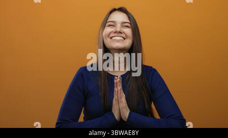 Die hispanische junge Frau lächelt mit gefalteten Händen vor isoliertem orangen Hintergrund, trägt einen blauen Pullover in fröhlichem Ausdruck, der Glück und Glück zum Ausdruck bringt Stockfoto