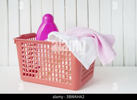 Weiße und rosafarbene Handtücher und Waschmittel in rosa Flasche befinden sich in einem roten Wäschekorb aus Plastik auf weißem hölzernem Hintergrund Stockfoto