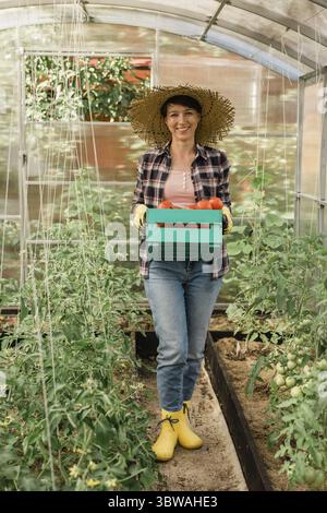 Eine Bio-Landwirtin, die eine Kiste voller frischer Produkte auf ihrem Hof hält. Glückliche junge Frau, die vor der Kamera lächelt, während sie in ihrem Gemüsegarten steht. Erfolgreich Stockfoto