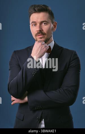 Junge Geschäftsleute in klassischem schwarzen Anzug, weißem Hemd und roter Krawatte. Gefaltet seine Hände, posiert vor einem blauen Studiohintergrund. Karriere, Reichtum, Busi Stockfoto