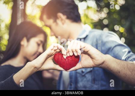 Zwei Hände der jungen Liebenden oder Paar halten roten Herzen Garn in Mitte an natürliche Outdoor Hintergrund für Valentines Tag. Urlaub und Konzept entspannen. Hone Stockfoto