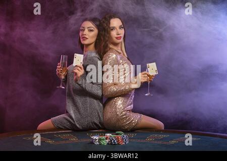 Zwei schöne Brünette-Damen spielen Poker im Casino, halten Gläser Champagner und Karten und sitzen auf dem Tisch mit Stapeln Chips drauf. Schwarz Stockfoto