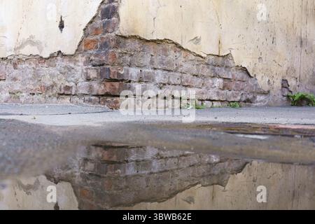 Eine Ziegelmauer mit einem Riss darin und eine Wasserpfütze, die die Wand reflektiert. Die Reflexion der Wand ist verzerrt und verschwommen, wodurch der Eindruck entsteht Stockfoto