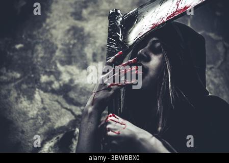 Hexe leckt Blut an den Fingern. Weiblicher Dämonenenengel in schwarzer Kleidung und Kapuze auf Grunge-Wand-Hintergrund. Halloween-Tag und Mystery-Konzept. Fantasie von Stockfoto